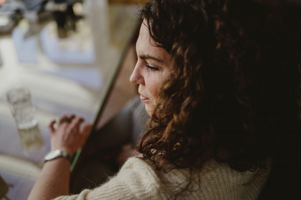 Esther Fischer, Mentorin und Therapeutin mit Fokus auf Nervensystemregulation, sitzt an einem Glastisch in einem lichtdurchfluteten Raum. Ihr Blick ist nachdenklich und nach innen gerichtet – ein Moment stiller Präsenz. Die Szene wirkt intim und zugleich kraftvoll: Ihre lockigen Haare fallen weich über die Schulter, während ihre Hände ruhig auf dem Tisch ruhen. Neben ihr ein Glas Wasser – Sinnbild für Klarheit und Erdung. Dieses Bild fängt die Essenz ihrer Arbeit ein: Menschen darin zu begleiten, aus innerer Anspannung in eine neue Form von Selbstverbundenheit zu finden. Als Mentorin für Selbstständige steht sie für eine tiefe, verkörperte Transformation, die es ermöglicht, nicht nur sichtbar, sondern auch wirksam zu wachsen – von innen nach außen.