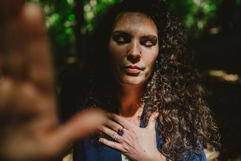 Esther Fischer steht mit geschlossenen Augen im lichten Wald, ihre rechte Hand liegt auf ihrem Herzen. Die warmen Sonnenstrahlen brechen durch das grüne Blätterdach und tauchen ihr Gesicht in natürliches Licht und Schatten. Die Farbfotografie verstärkt die Atmosphäre von Erdung, Präsenz und innerer Verbundenheit. Als Mentorin bei wirksam wachsen begleitet Esther Fischer Frauen in Verantwortung durch Nervensystemarbeit, somatisches Mentoring und bewusste Transformation – getragen von Tiefe, Achtsamkeit und therapeutischer Klarheit.