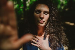 Esther Fischer steht mit geschlossenen Augen im lichten Wald, ihre rechte Hand liegt auf ihrem Herzen. Die warmen Sonnenstrahlen brechen durch das grüne Blätterdach und tauchen ihr Gesicht in natürliches Licht und Schatten. Die Farbfotografie verstärkt die Atmosphäre von Erdung, Präsenz und innerer Verbundenheit. Als Mentorin bei wirksam wachsen begleitet Esther Fischer Frauen in Verantwortung durch Nervensystemarbeit, somatisches Mentoring und bewusste Transformation – getragen von Tiefe, Achtsamkeit und therapeutischer Klarheit.