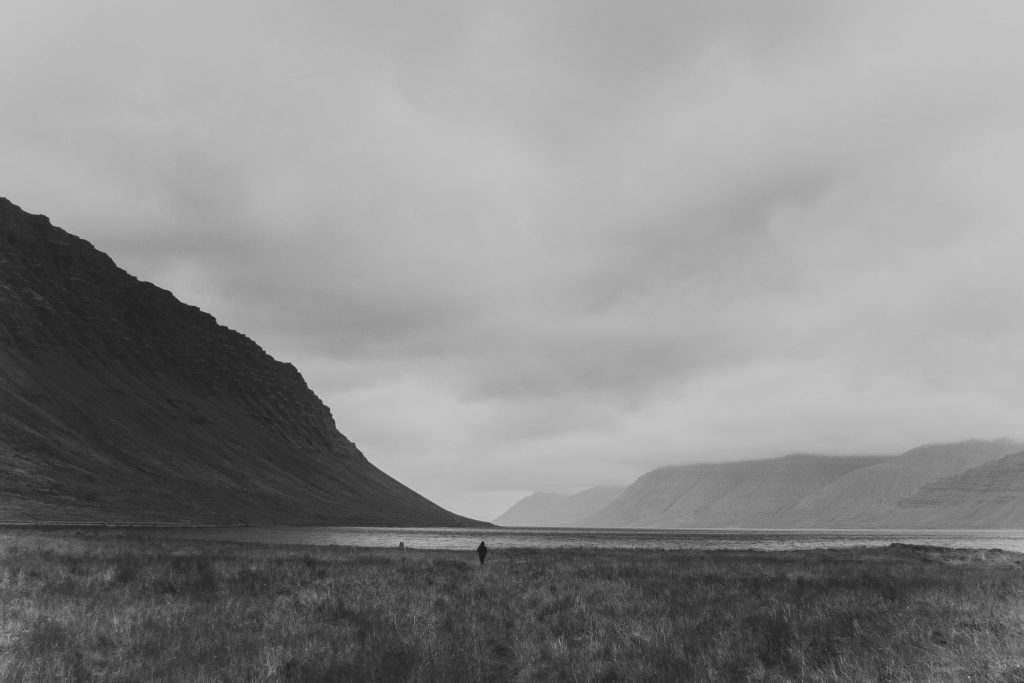 Ein schwarz-weißes Landschaftsfoto zeigt eine weitläufige, grasbewachsene Ebene zwischen dunklen Berghängen. In der Mitte des Bildes steht eine einzelne, kleine menschliche Figur am Ufer eines ruhigen Gewässers, das sich in die Ferne zwischen die Berge zieht. Der Himmel ist wolkenverhangen, die Szene wirkt ruhig, karg und introspektiv. Das Bild transportiert ein Gefühl von Einsamkeit, Weite und innerer Einkehr – fast meditativ.