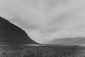 Ein schwarz-weißes Landschaftsfoto zeigt eine weitläufige, grasbewachsene Ebene zwischen dunklen Berghängen. In der Mitte des Bildes steht eine einzelne, kleine menschliche Figur am Ufer eines ruhigen Gewässers, das sich in die Ferne zwischen die Berge zieht. Der Himmel ist wolkenverhangen, die Szene wirkt ruhig, karg und introspektiv. Das Bild transportiert ein Gefühl von Einsamkeit, Weite und innerer Einkehr – fast meditativ.