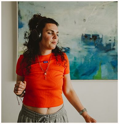 Esther Fischer steht vor einem abstrakten, blaugrünen Gemälde und trägt ein auffällig orangenes Shirt sowie Kopfhörer. Ihre Augen sind geschlossen, der Körper leicht in Bewegung – ein Moment ganz im Gefühl, frei und verbunden. Ihre Haltung und ihr Gesichtsausdruck spiegeln Körperbewusstsein und Präsenz wider. Als Mentorin für somatische Selbstführung bei wirksam wachsen nutzt sie u. a. Musik und Bewegung, um das Nervensystem zu regulieren und emotionale Prozesse bewusst zu begleiten – besonders für Menschen in Führung und Wandel.