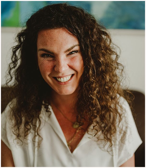 Esther Fischer sitzt auf einem dunklen Sofa vor einem abstrakten, blau-grünen Kunstwerk. Sie trägt eine weiße Bluse, ihre lockigen Haare umrahmen ihr Gesicht, während sie direkt in die Kamera lächelt – offen, herzlich und voller Energie. Diese Nahaufnahme bringt ihre menschliche Tiefe und Zugänglichkeit zum Ausdruck. Als Mentorin bei wirksam wachsen unterstützt sie Menschen in Führung und Entwicklung mit Nervensystemarbeit, emotionaler Klarheit und therapeutischer Begleitung auf Augenhöhe.