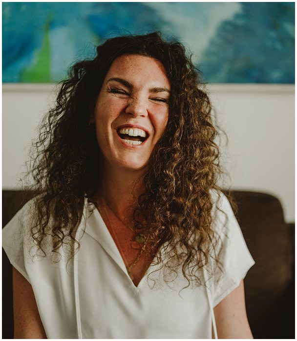 Esther Fischer sitzt auf einem dunklen Sofa vor einem abstrakten, meeresfarbenen Kunstwerk. Sie trägt ein weißes Shirt, ihre Locken fallen offen über die Schultern. Mit einem offenen, kraftvollen Lachen blickt sie leicht zur Seite – ein authentischer Moment voller Lebendigkeit und Vertrauen. Als Mentorin und Therapeutin bei wirksam wachsen begleitet sie Führungskräfte durch Nervensystemarbeit, somatisches Mentoring und emotionale Klarheit.