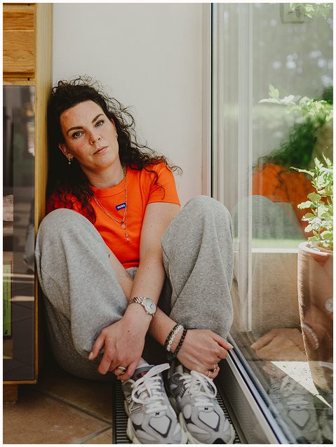 Esther Fischer sitzt auf dem Boden vor einer großen Fensterscheibe, die Beine angezogen, der Blick offen und nachdenklich. Sie trägt ein leuchtend oranges Shirt und graue Jogginghose, ihr Körper wirkt entspannt, der Ausdruck ruhig und präsent. Durch das Fenster fällt sanftes Licht auf ihre Seite. Als Mentorin bei wirksam wachsen steht sie hier sinnbildlich für authentische Selbstbegegnung, innere Klärung und Nervensystemarbeit für Frauen, die sich in anspruchsvollen Rollen selbst nicht verlieren wollen.