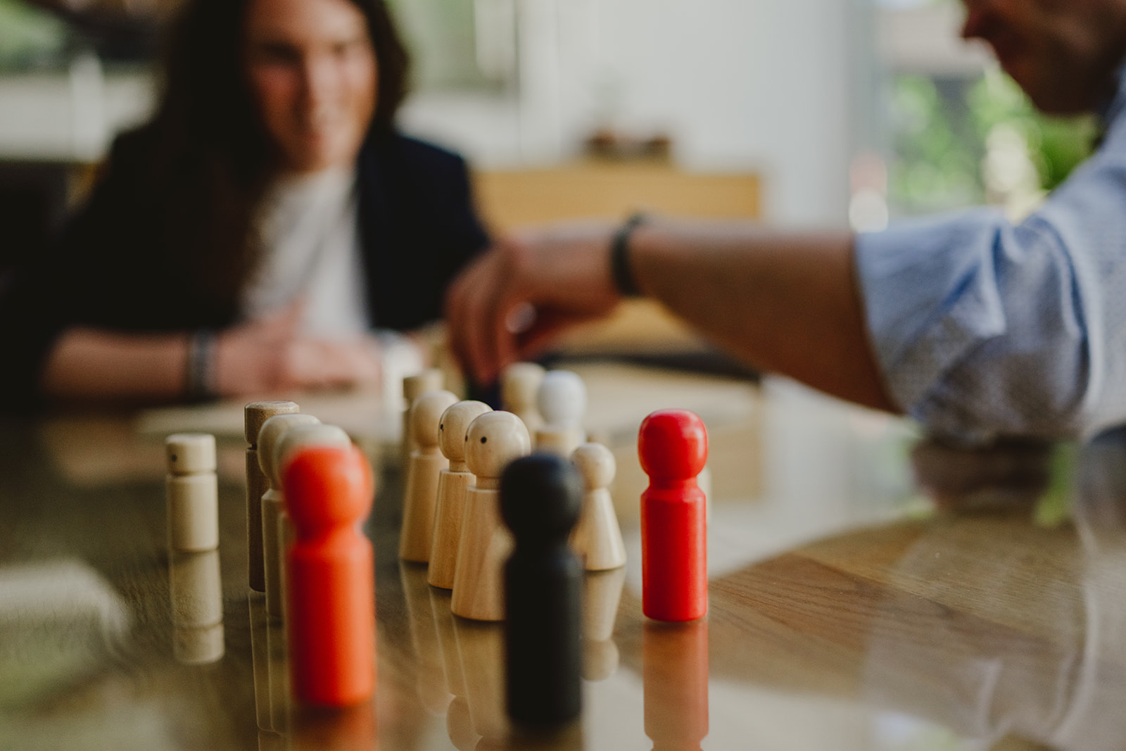 Holzfiguren verschiedener Formen und Farben stehen auf einem Tisch in strukturierter Anordnung – eine typische Szene aus der systemischen Aufstellungsarbeit. Im unscharfen Hintergrund erkennt man zwei Personen, darunter Esther Fischer, im Gespräch vertieft. Die schwarz-weiße Aufnahme lenkt den Fokus auf die Dynamik im Setting. Als Mentorin für systemische Prozessbegleitung und Nervensystemarbeit bei wirksam wachsen nutzt Esther Fischer Aufstellungen, um unbewusste Muster sichtbar und transformierbar zu machen – insbesondere für Führungskräfte und Selbstständige mit Wunsch nach tiefer Klärung.