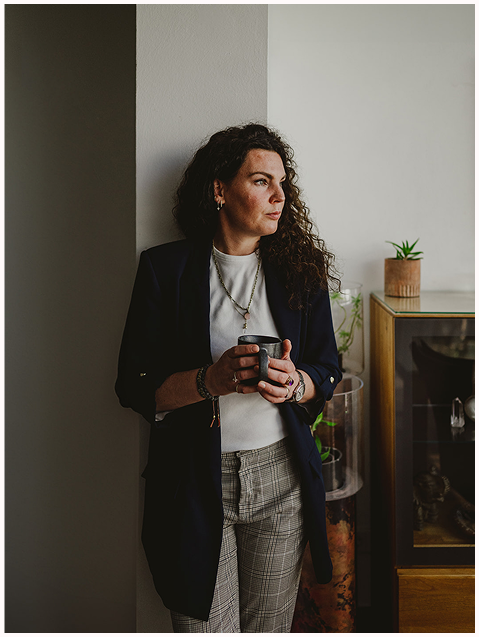 Esther Fischer lehnt mit einer dunklen Tasse in der Hand an einer weißen Wand, der Blick ruhig und nachdenklich zum Fenster gerichtet. Sie trägt eine karierte Hose, ein weißes Shirt und einen dunklen Blazer. Im Hintergrund stehen Zimmerpflanzen und natürliche Deko-Elemente. Dieses Bild strahlt Ruhe, Reflexion und innere Präsenz aus – Qualitäten, die sie in ihrer Arbeit als Mentorin bei wirksam wachsen verkörpert. Ihre Begleitung in Nervensystemarbeit, therapeutischem Mentoring und bewusster Selbstführung schafft Raum für Klarheit, Regeneration und tiefe Veränderung.