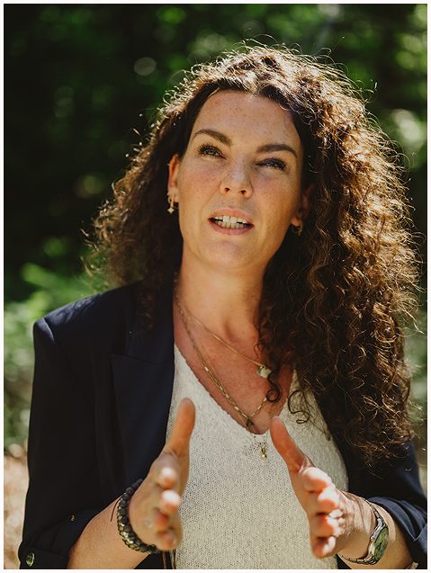 Esther Fischer steht inmitten eines lichten Waldes, ihre Hände sind in einer erklärenden Geste geöffnet, der Gesichtsausdruck lebendig und konzentriert. Ihr Blick ist leicht erhoben, sie spricht mit Nachdruck und gleichzeitig Ruhe. Die Sonnenstrahlen akzentuieren ihre lockigen Haare, sie trägt ein dunkles Jackett über einem weißen Top mit strukturierter Oberfläche.