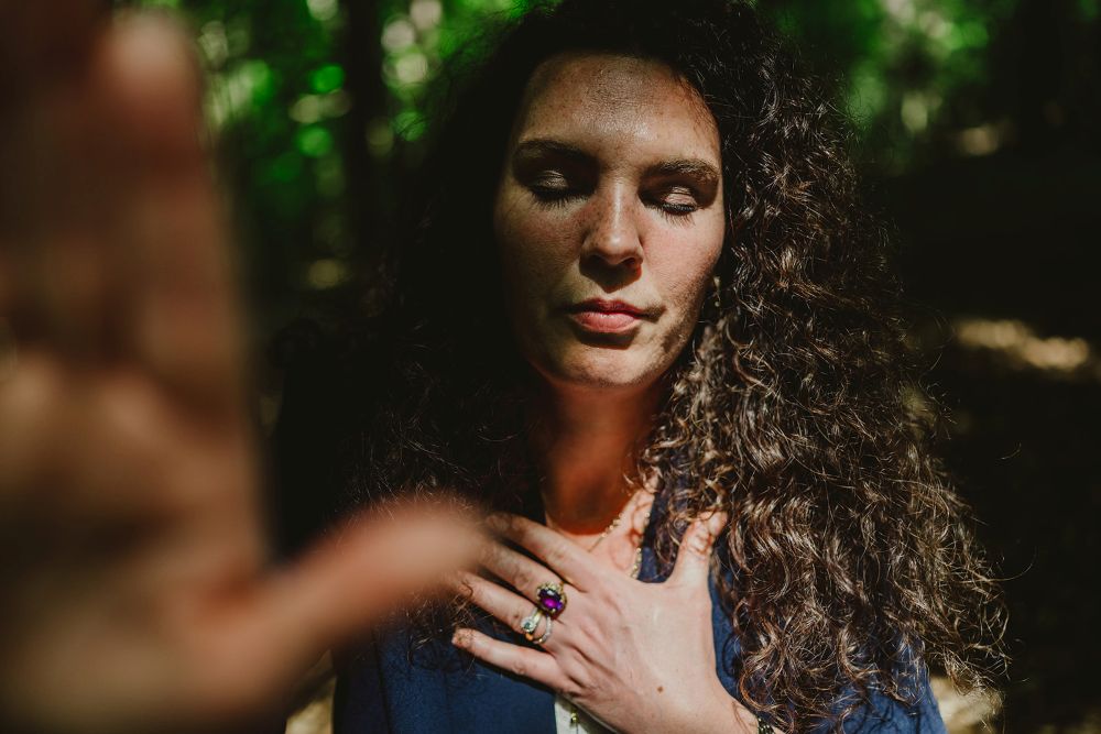 Esther Fischer steht mit geschlossenen Augen im lichten Wald, ihre rechte Hand liegt auf ihrem Herzen. Die warmen Sonnenstrahlen brechen durch das grüne Blätterdach und tauchen ihr Gesicht in natürliches Licht und Schatten. Die Farbfotografie verstärkt die Atmosphäre von Erdung, Präsenz und innerer Verbundenheit. Als Mentorin bei wirksam wachsen begleitet Esther Fischer Frauen in Verantwortung durch Nervensystemarbeit, somatisches Mentoring und bewusste Transformation – getragen von Tiefe, Achtsamkeit und therapeutischer Klarheit.