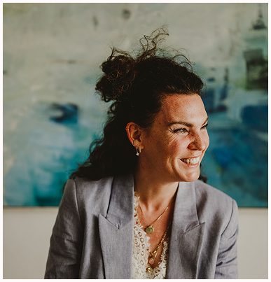 Esther Fischer sitzt vor einem abstrakten Bild in Meeresfarben, ihr Blick geht strahlend zur Seite. Sie trägt einen hellvioletten Blazer und eine bestickte Bluse, ihre Haare sind locker hochgesteckt. Ein Moment voller Lebensfreude, Wärme und natürlicher Präsenz. Als Mentorin bei wirksam wachsen steht sie für empathische Begleitung, emotionale Klarheit und somatische Selbstführung – speziell für Frauen, die sich in beruflichen Rollen mit Tiefe und Verantwortung neu ausrichten wollen.