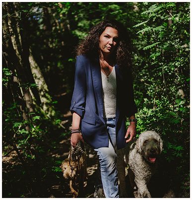 Esther Fischer geht mit zwei Hunden durch einen sonnendurchfluteten Waldweg. Sie trägt einen dunklen Blazer, ein helles Top und Jeans – ein starker Kontrast zur lebendigen Natur um sie herum. Licht und Schatten spielen auf ihrem Gesicht, ihre Ausstrahlung wirkt ruhig, fokussiert und verbunden. Dieses Bild steht symbolisch für ihre Haltung bei wirksam wachsen: Mentoring und Nervensystemarbeit im Einklang mit Natur, Körper und Klarheit.
