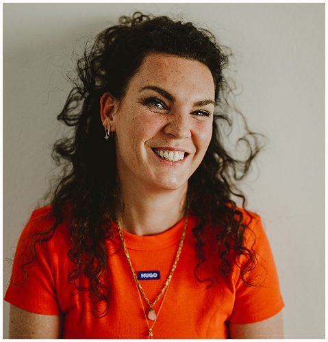 Esther Fischer steht vor einer hellen Wand und trägt ein leuchtend orangenes Shirt mit dem Schriftzug „HUGO“. Ihre offenen Locken rahmen ihr Gesicht, sie lacht herzlich in die Kamera. Zwei goldene Ketten und ein Stern-Ohrring unterstreichen ihre individuelle Ausstrahlung. Dieses Porträt zeigt die Mentorin für emotionale Gesundheit, Therapie und Führungskräfteentwicklung bei wirksam wachsen von ihrer lebendigen, authentischen Seite – sichtbar nahbar und gleichzeitig klar positioniert.