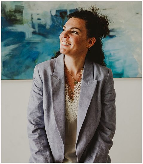 Esther Fischer steht vor einem großformatigen, abstrakten Kunstwerk in Blau- und Grüntönen. Sie trägt einen hellen Blazer und eine Spitzenbluse, ihr Blick geht strahlend zur Seite, das Lächeln offen und herzlich. Dieses Bild wirkt zugleich professionell und nahbar – es verkörpert ihre Haltung als Mentorin für Selbstführung, Nervensystemarbeit und Führungskräfteentwicklung bei wirksam wachsen. Ihre Arbeit verbindet Tiefe, Lebensfreude und echte Verbindung – sichtbar auch in ihrer Ausstrahlung.